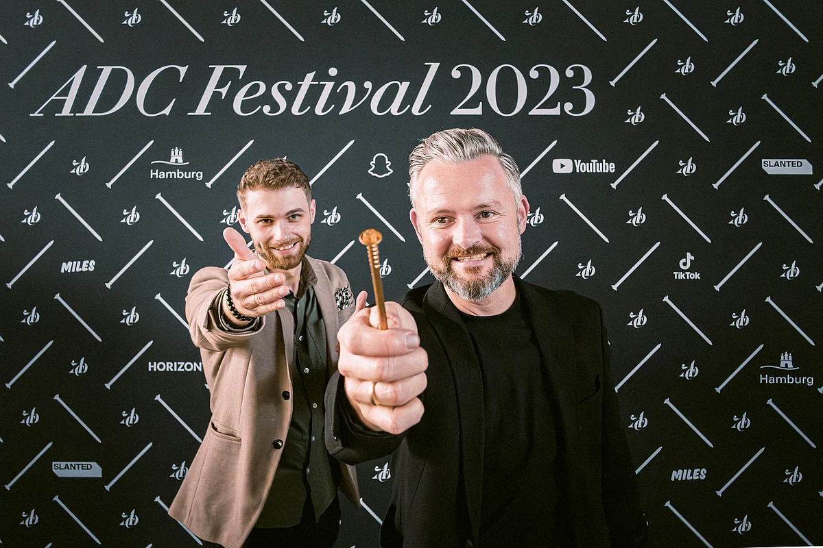 Männer posieren vor ADC Festival 2023 Text Zwei Männer posieren vor einem schwarzen Hintergrund mit dem Text "ADC Festival 2023" und halten einen Gegenstand in der Hand.