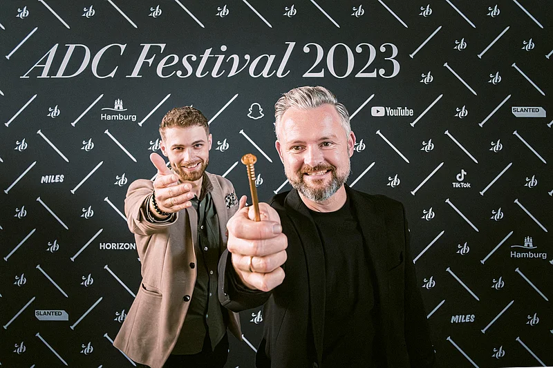 Zwei Männer posieren vor einem schwarzen Hintergrund mit dem Text "ADC Festival 2023" und halten einen Gegenstand in der Hand.