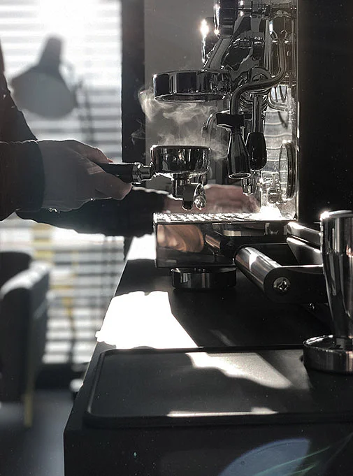 Kaffeezubereitung mit Siebträger Eine Hand hält einen Siebträger vor einer Kaffeemaschine, aus der Dampf aufsteigt.
