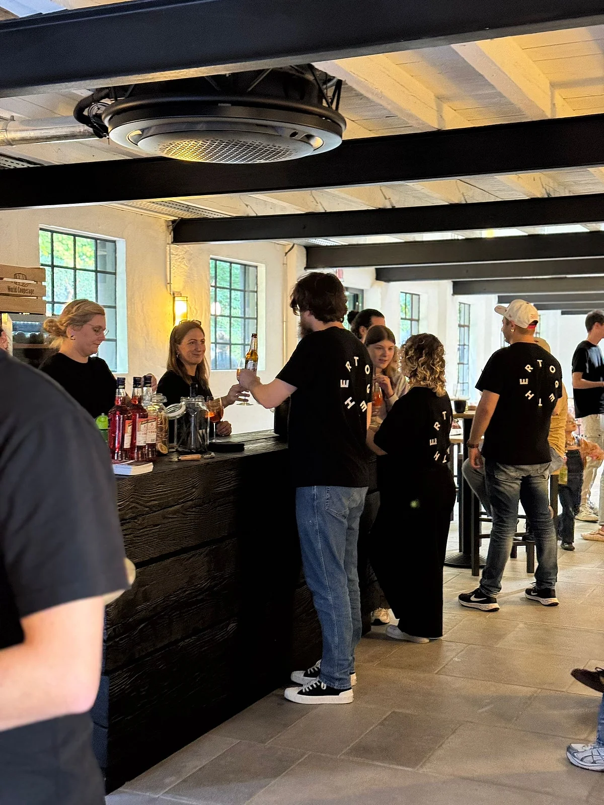 Bar mit Menschen in schwarzen T-Shirts Eine Bar mit Menschen in schwarzen T-Shirts, die Getränke servieren und konsumieren. Tische und Fenster sind sichtbar.